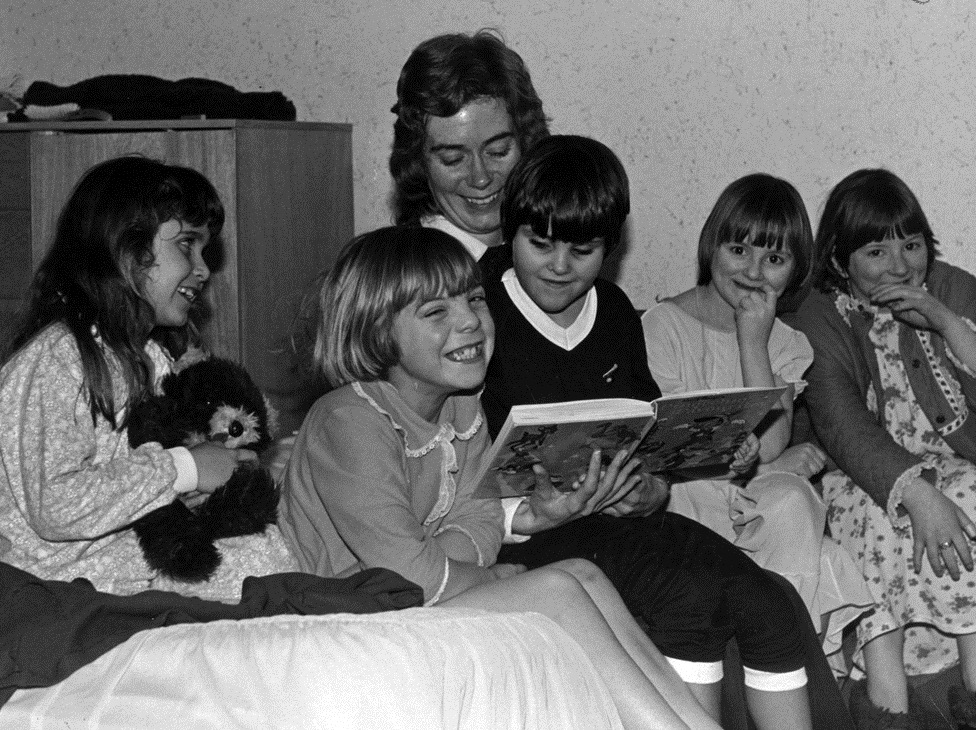 A black and white photograph of 5 young siblings sat with their mother. Several are laughing. One is reading a book. 