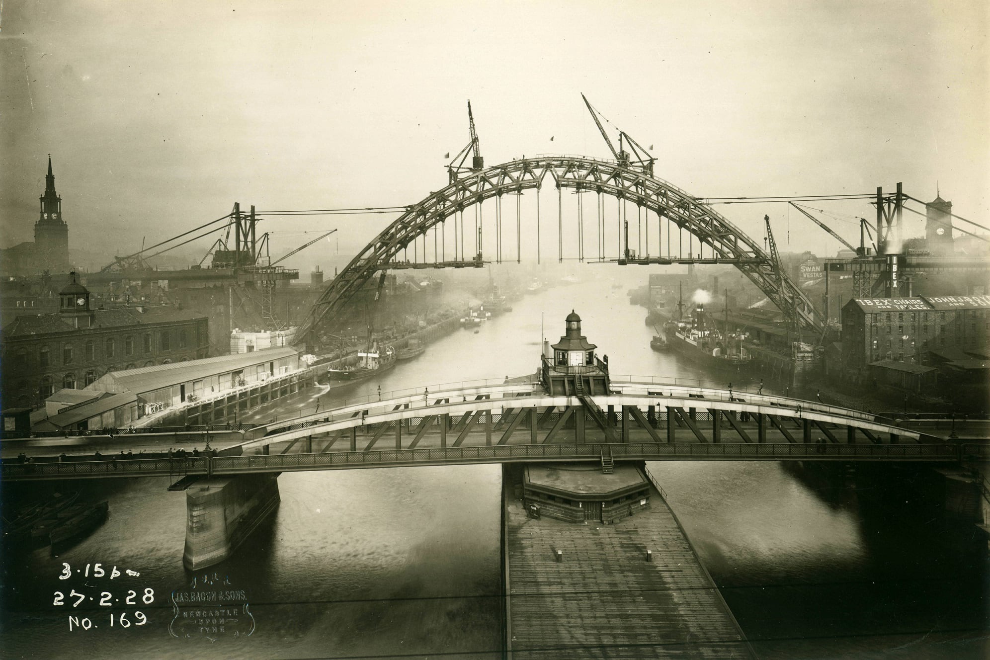 A black and white photograph stamped 3.15pm 27.2.28, looking down the Tyne, over the Swing Bridge to a half built Tyne bridge. The air is thick with smog. There are various steam boats docked on the sides of the river.